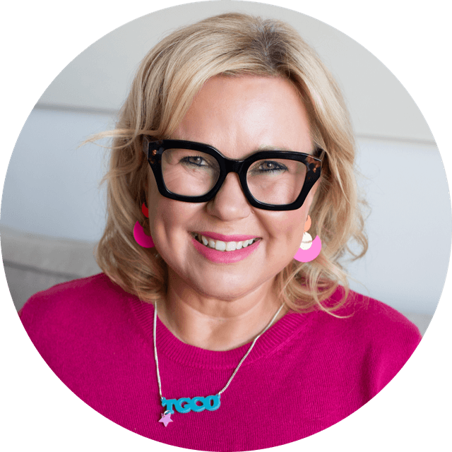 Close-up portrait of a smiling blonde woman wearing black-framed glasses, a pink sweater, pink earrings and a turquoise pendant necklace.