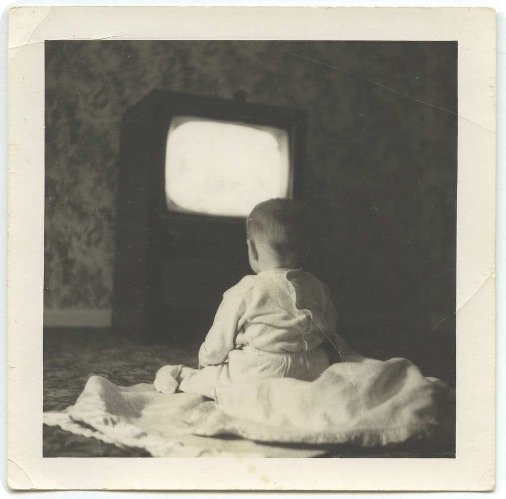 A baby sits on a blanket, watching an old-fashioned TV in a dimly lit room. The TV screen glows brightly in the background.