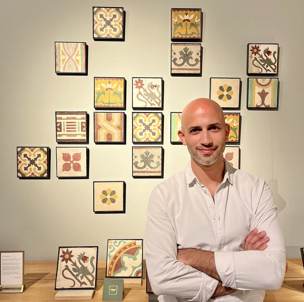 Man in white shirt stands with arms crossed before a wall displaying colorful, patterned tiles in a grid arrangement.