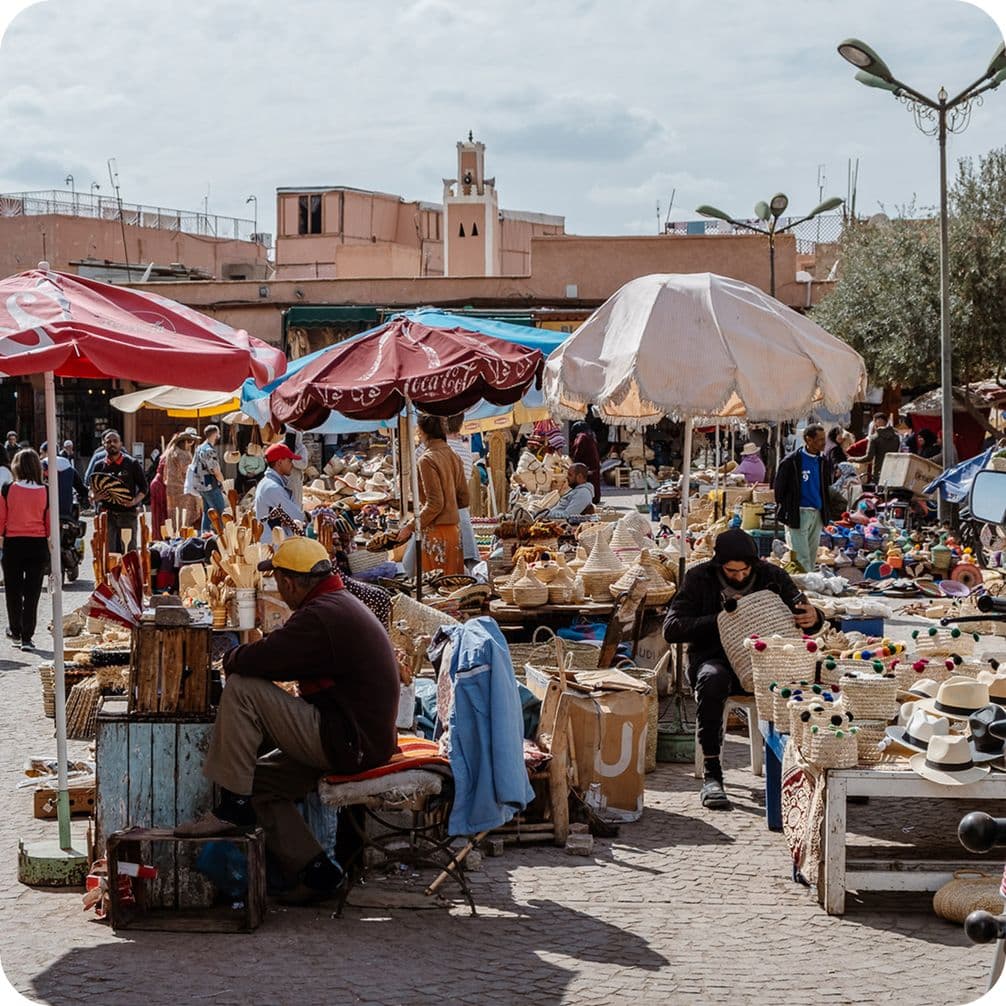 Bustling outdoor market with colorful umbrellas, vendors selling crafts and hats, and people walking around under a cloudy sky.