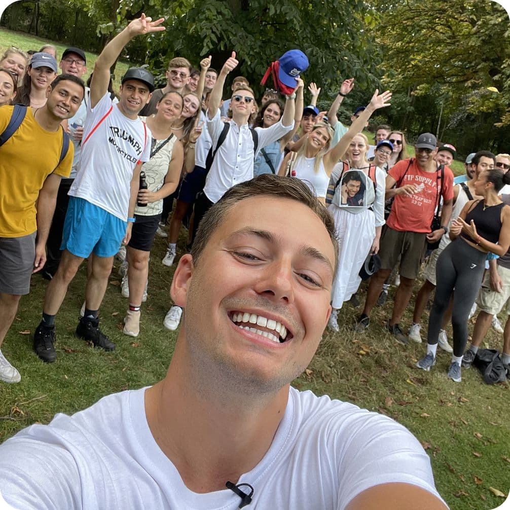 A smiling person takes a selfie with a large, cheerful group of people outdoors, surrounded by trees.