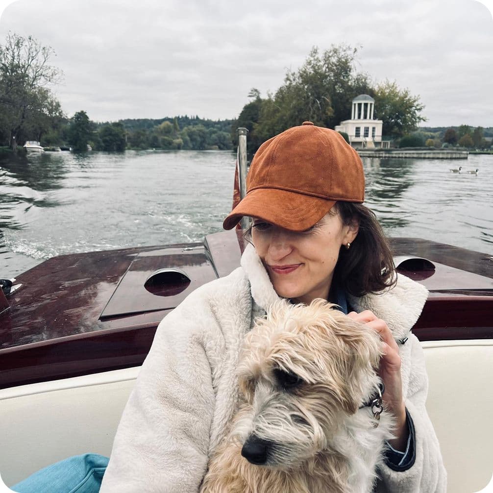 Woman in brown cap and cream jacket cuddling a small scruffy dog on a boat, river and boathouse in the background.