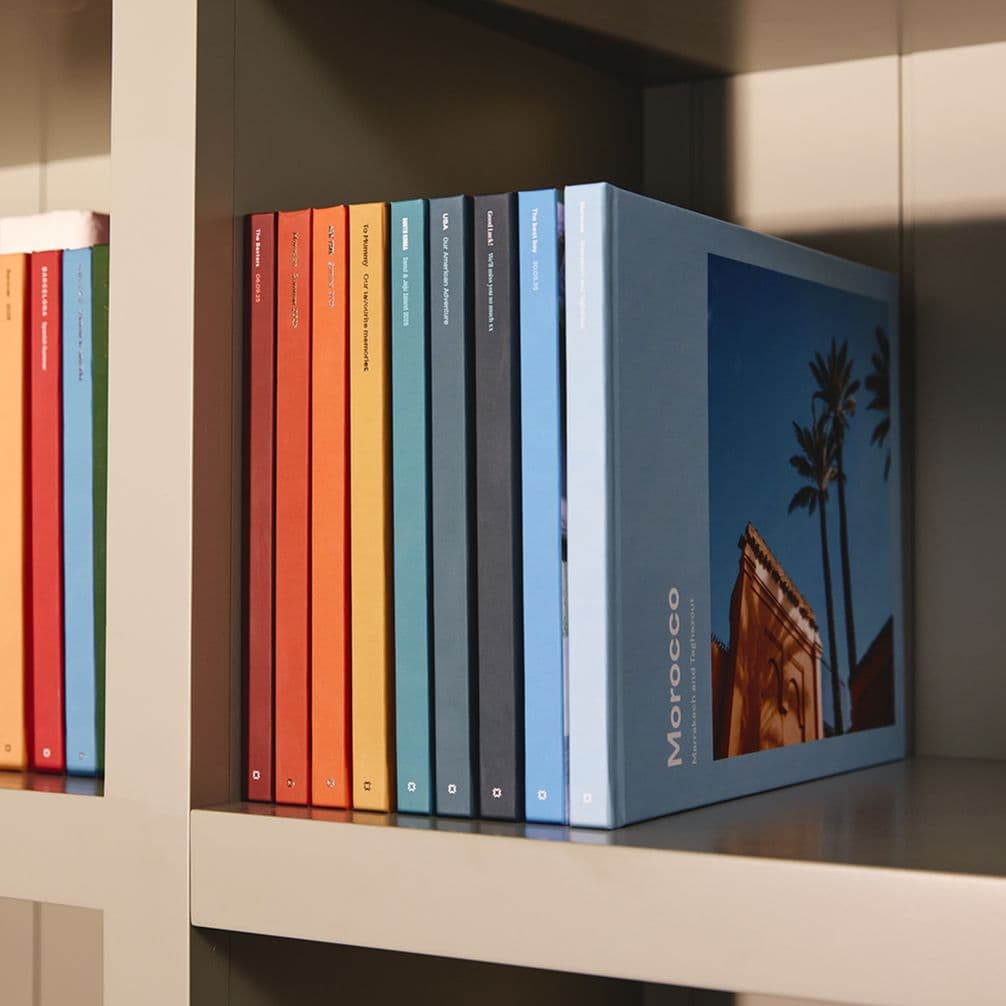 Shelf holding a row of colorful slim books beside a larger blue photo book titled Morocco showing palm trees and architecture.