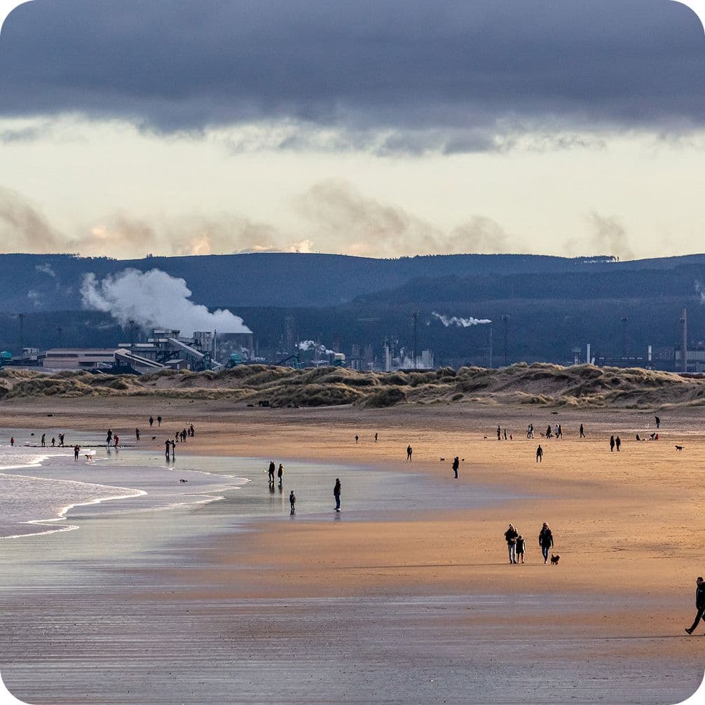 People walking on a wide, sandy beach with distant industrial buildings and smoke under a cloudy sky.