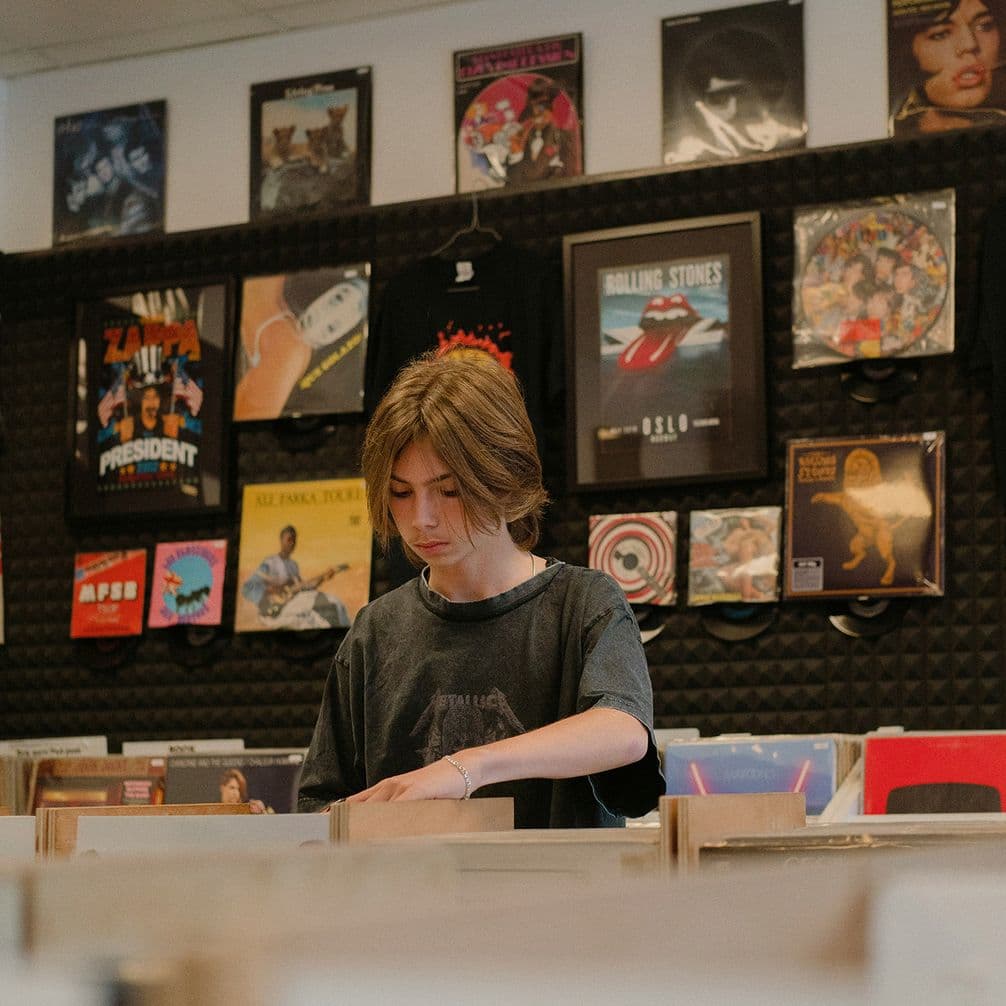 Person browsing vinyl records in a record store, flipping through crate bins beneath a wall of framed album covers.