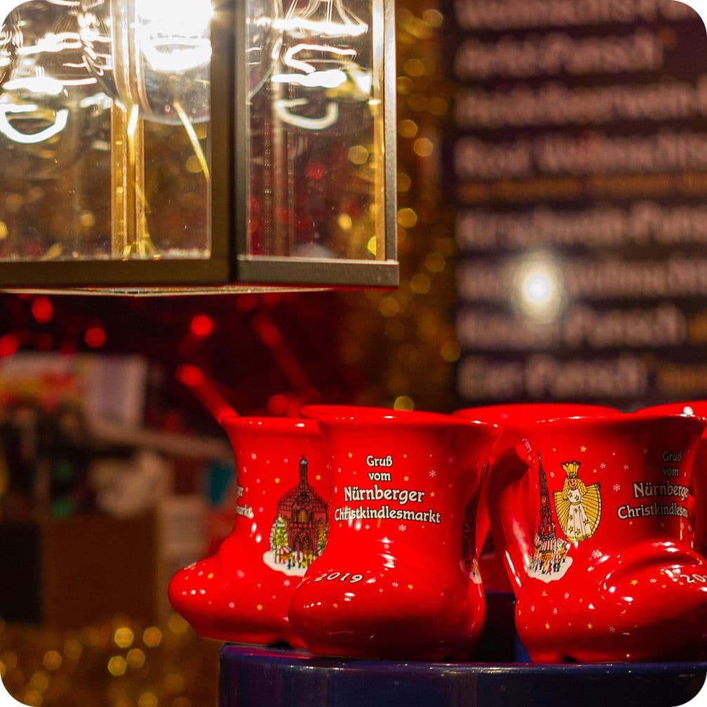 Red boot-shaped mugs on display at a festive market stall, with a glowing light above and a blurred background of text and golden decorations.