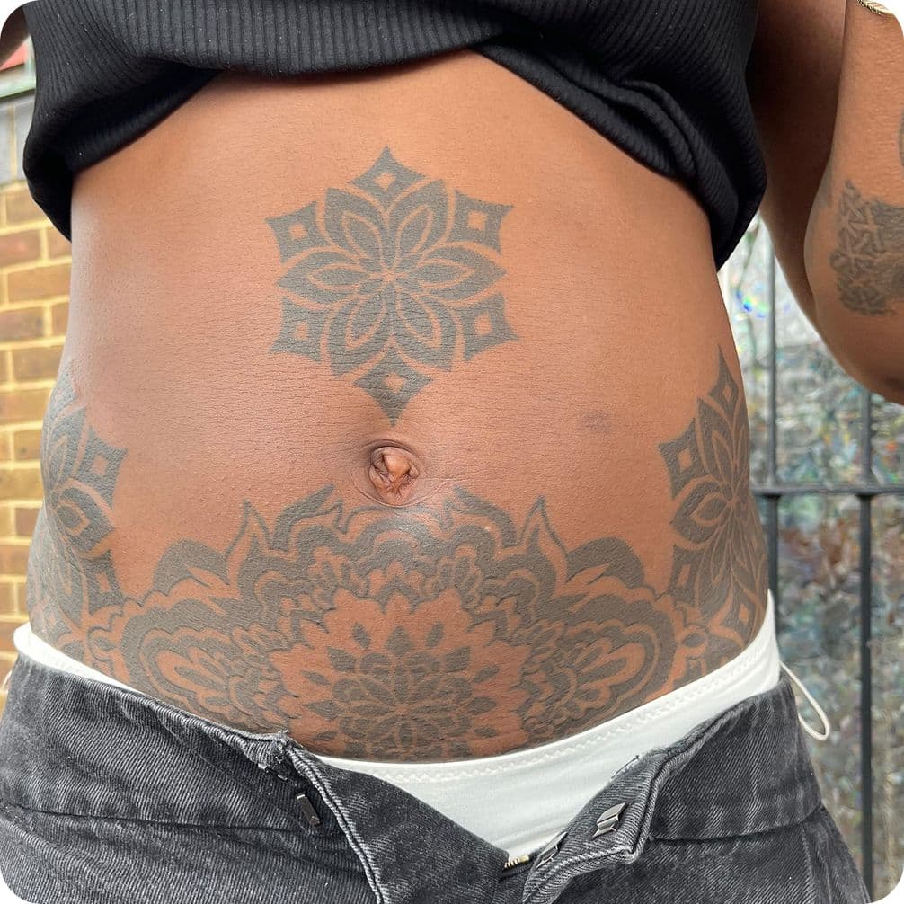 Close-up of a person's torso with intricate floral tattoos around the navel, wearing a black top and partially unzipped jeans.