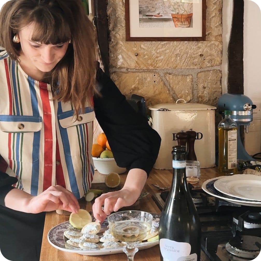 A woman in a striped vest prepares food in a rustic kitchen, with wine and citrus on the counter.