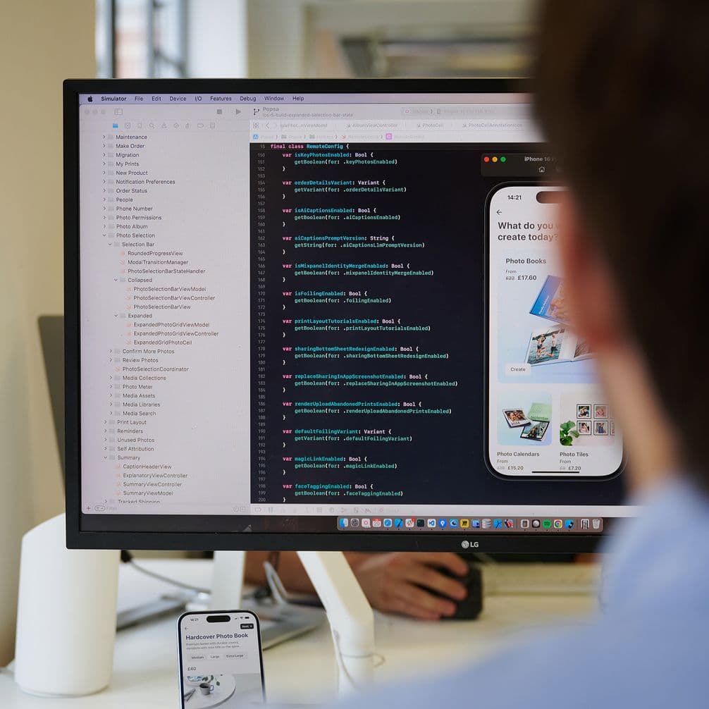 Person seated, viewing computer screen displaying code and mobile app design. iPhone with code rests nearby. Modern office setting.