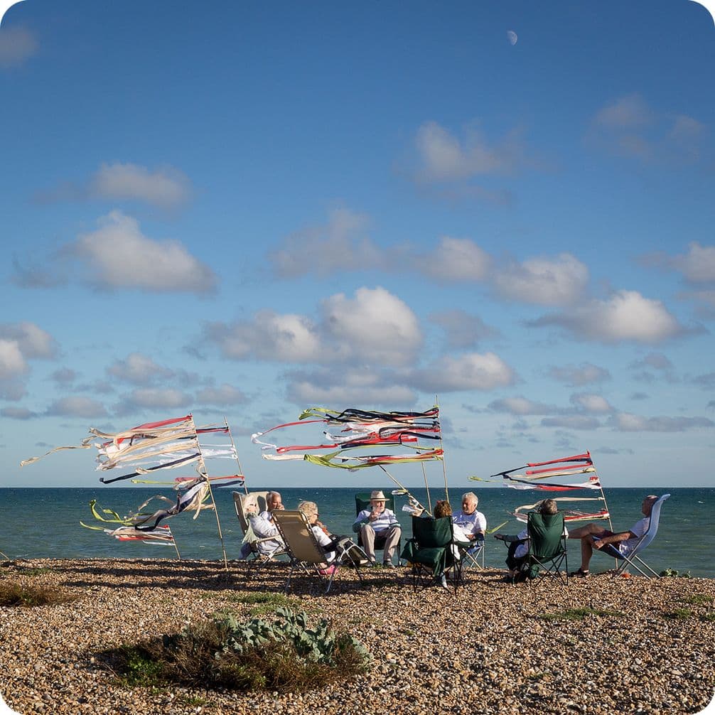 People sitting on a pebble beach with colorful flags flying, under a blue sky with scattered clouds and a visible horizon.