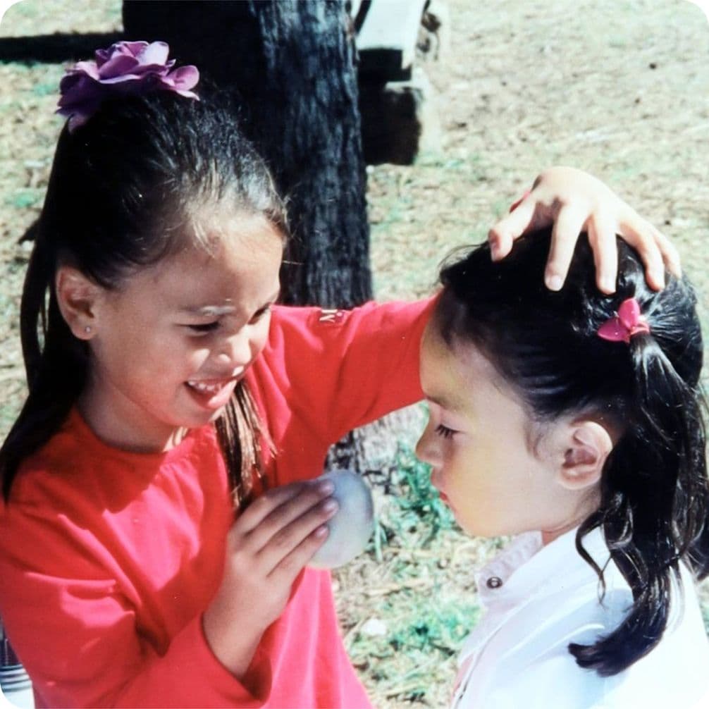 Two young girls outdoors, one in a red shirt holding a balloon near the other's face. Both have flowers in their hair.