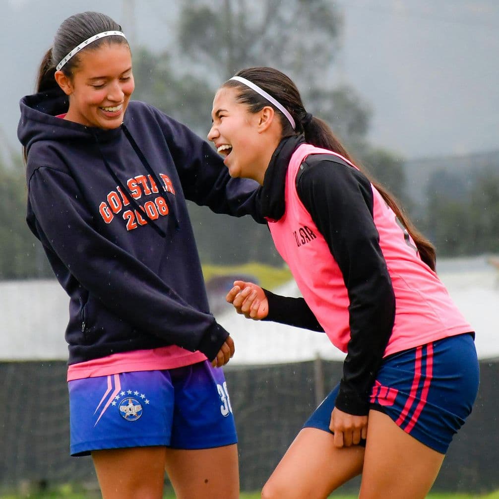 Two teenage soccer players in sportswear laughing together on a rainy field, one in a dark hoodie, one in a pink training vest.