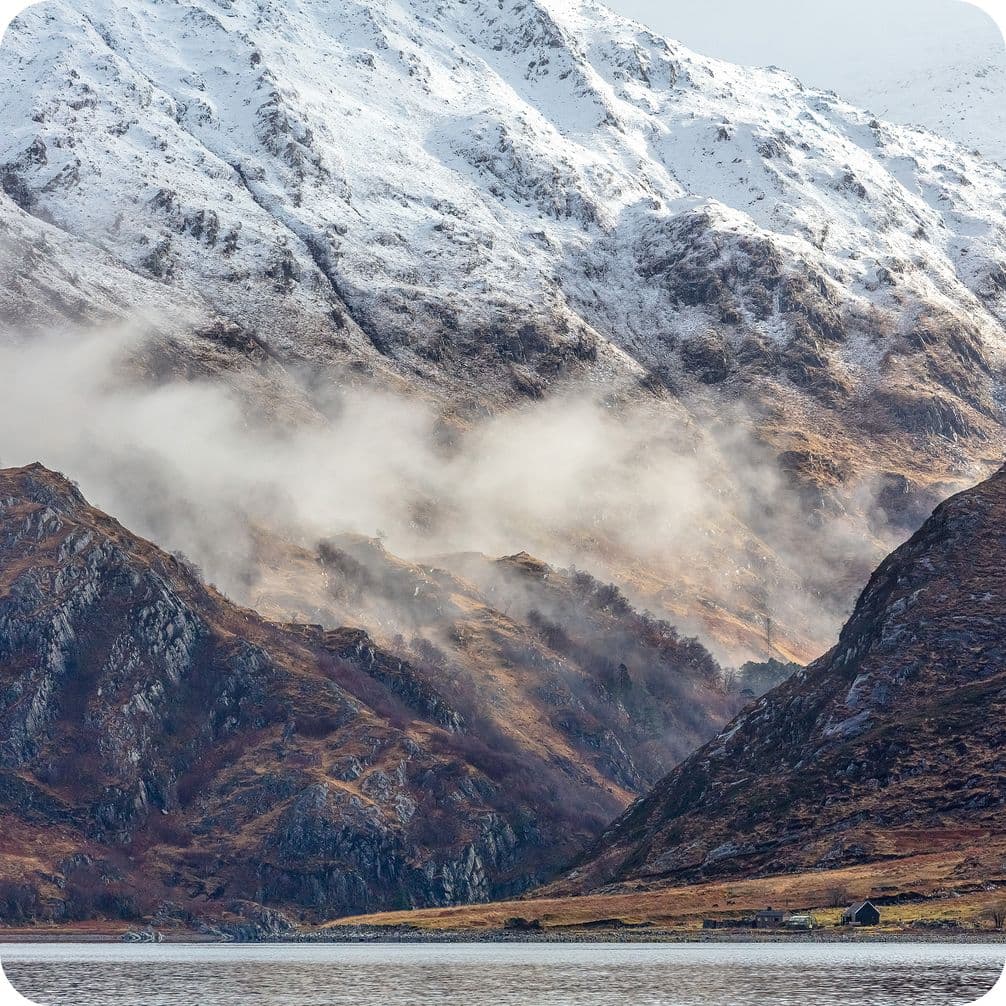 Snow-capped mountains with misty clouds, rocky slopes, and a calm lake in the foreground, creating a serene landscape.