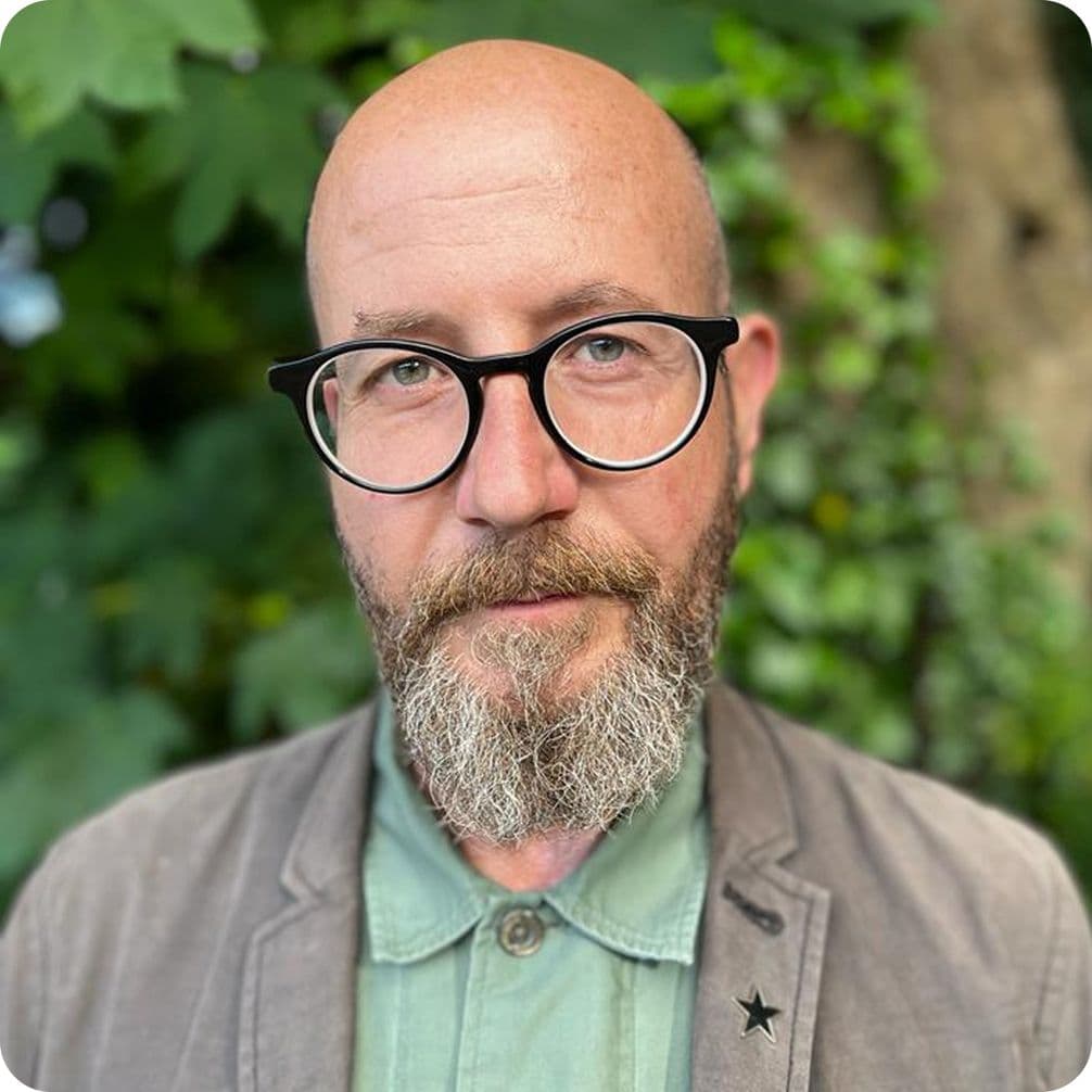 Bald man with round black glasses and gray beard, wearing a light green shirt and gray jacket, blurred leafy background.