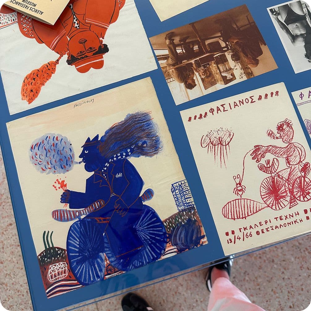 A display of colorful, artistic posters on a blue table, featuring abstract figures on bicycles and handwritten text in Greek.