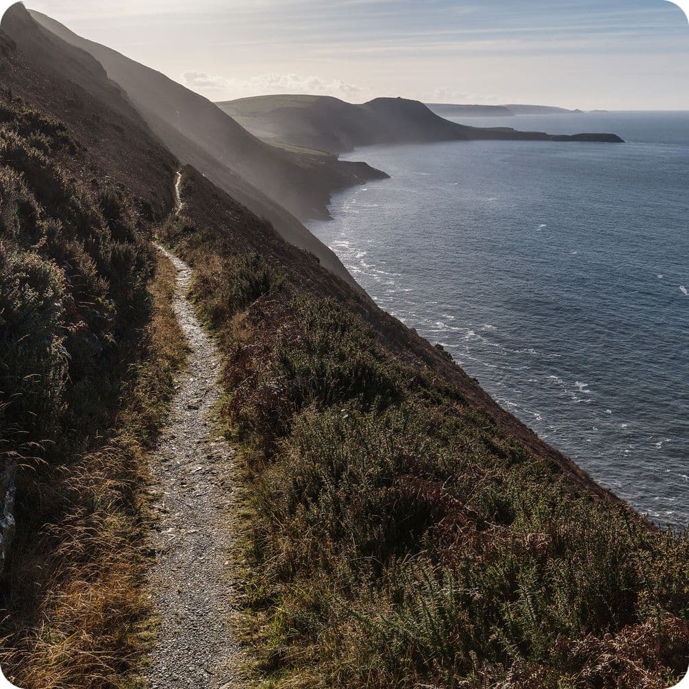Coastal cliffside path with rugged terrain, overlooking a vast ocean under a clear sky, distant hills visible across the water.