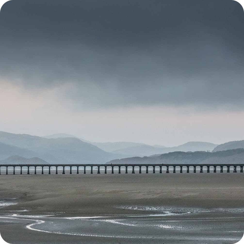 A long, narrow bridge stretches across a vast, empty landscape with distant hills under a moody, overcast sky.