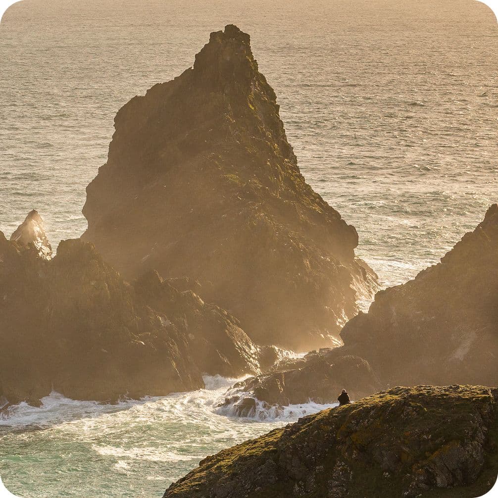 Jagged sea stacks rise from the ocean, surrounded by mist and waves, with a lone figure sitting on a grassy cliff in the foreground.