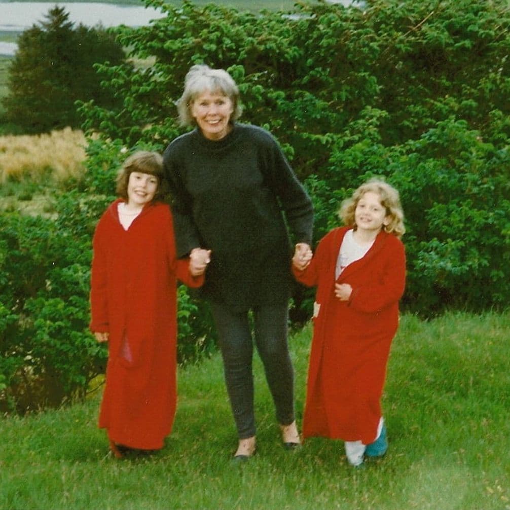 A smiling woman holds hands with two children wearing red robes, standing on grass with lush greenery in the background.