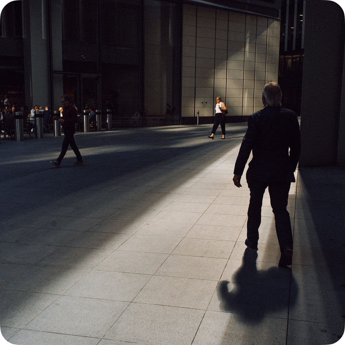People walking on a sunlit urban sidewalk, with long shadows cast by tall buildings.