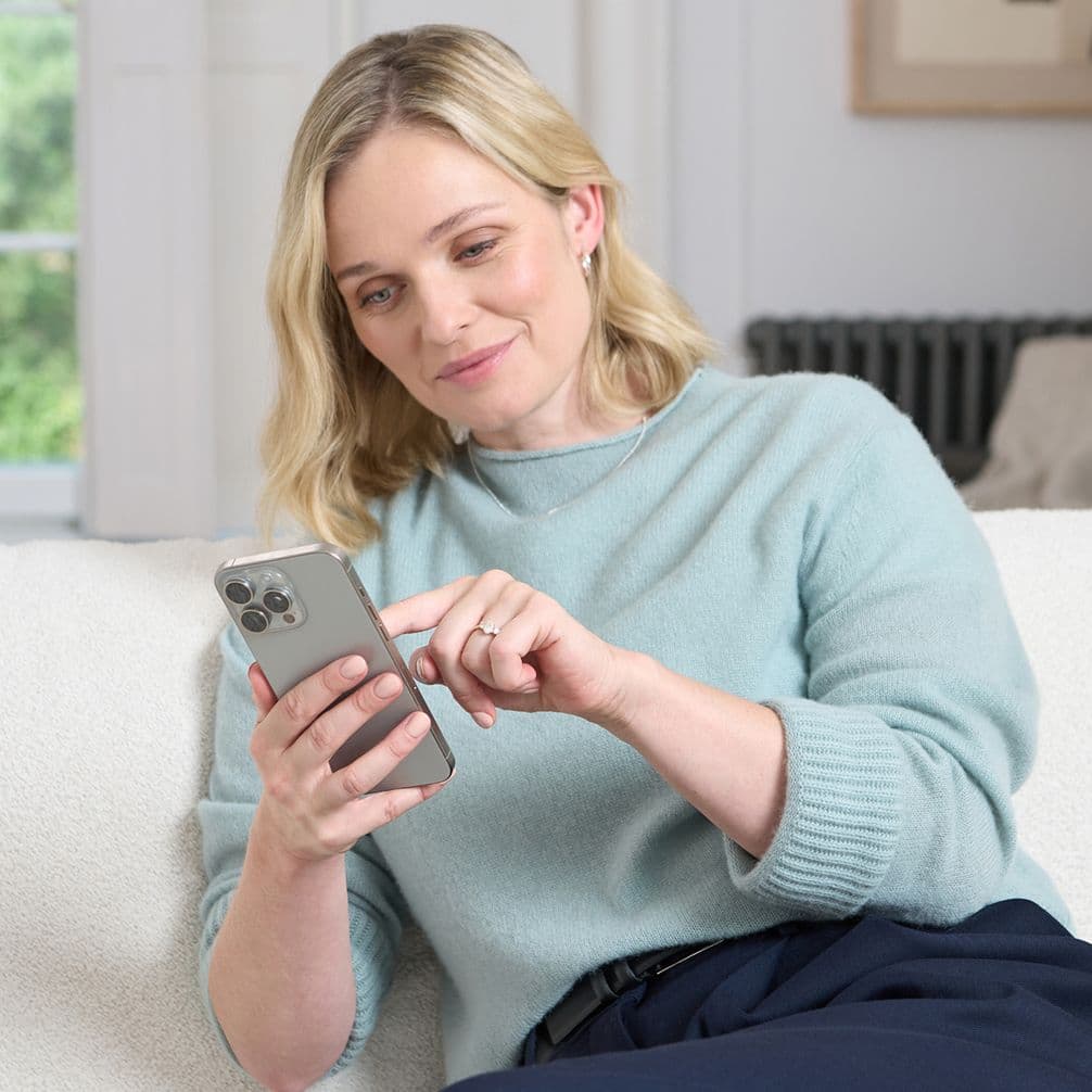 Blonde woman in a light blue sweater reclining on a sofa, smiling as she taps a smartphone screen.
