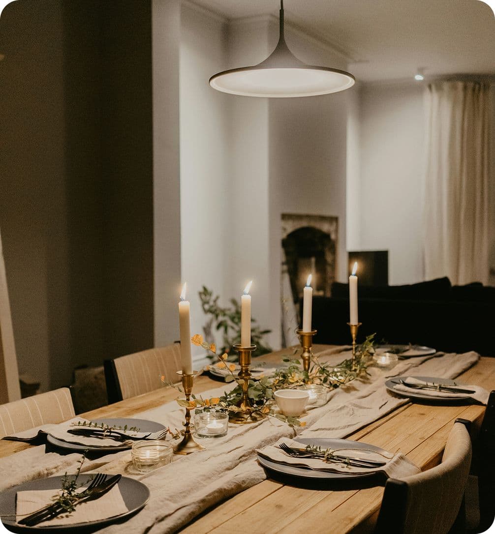 A warmly lit dining table set for six with candles, plates, and cutlery, adorned with greenery and a soft table runner.