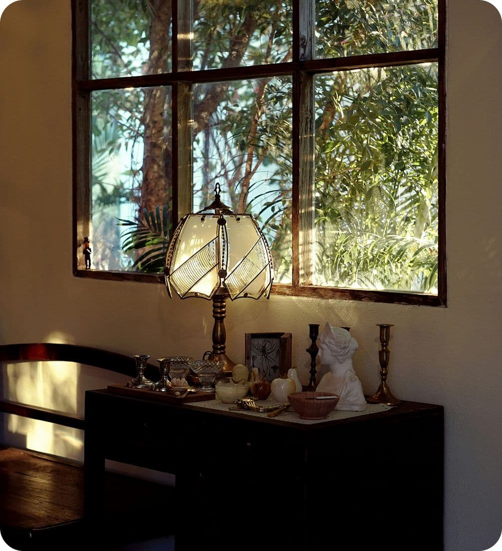 A cozy room with a vintage lamp on a wooden table, decorative items, and a window showing sunlight filtering through trees outside.
