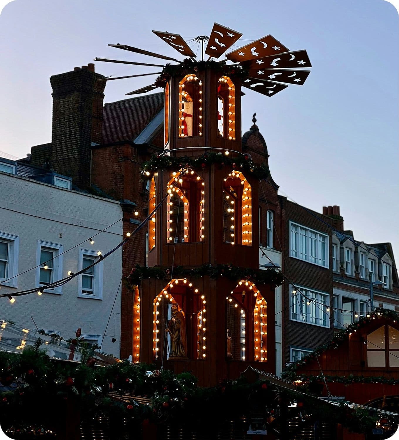 Three-tier wooden Christmas market pyramid with lit arches and rotating star-shaped blades, garlands and string lights against dusk buildings.