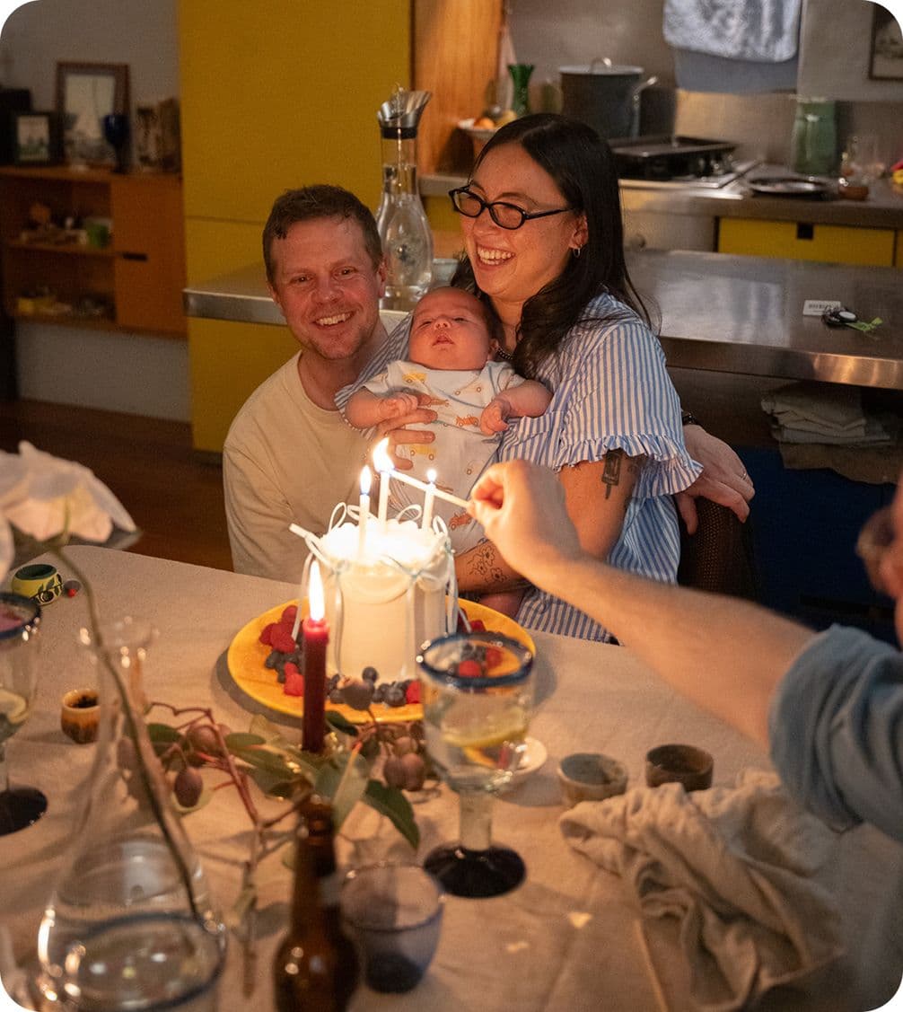 A couple smiles while holding a baby in front of a lit birthday cake. Candles and tableware are arranged on the table.