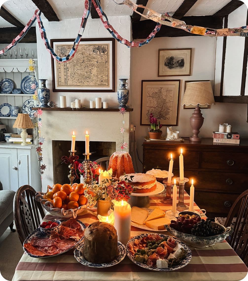 Cozy dining room with a festive table spread, featuring candles, fruit, meats, and cakes, adorned with paper chains and elegant decor.