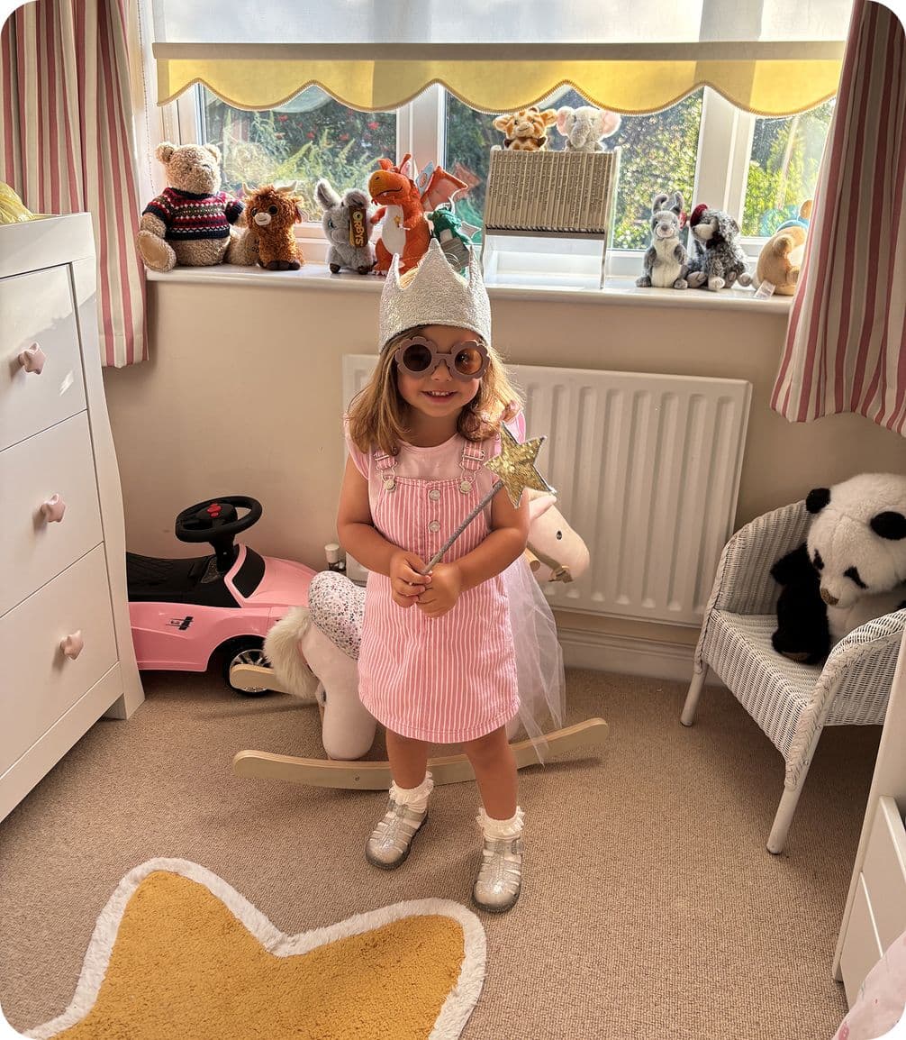 A child in a pink dress and crown holds a wand in a toy-filled room, with stuffed animals and a toy car in the background.
