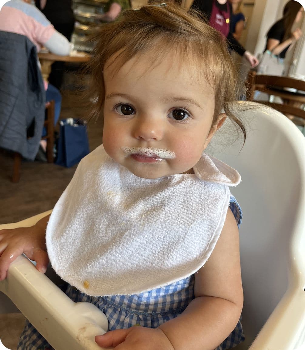 A baby with a milk mustache sits in a high chair, wearing a white bib and blue checkered outfit, looking curiously at the camera.