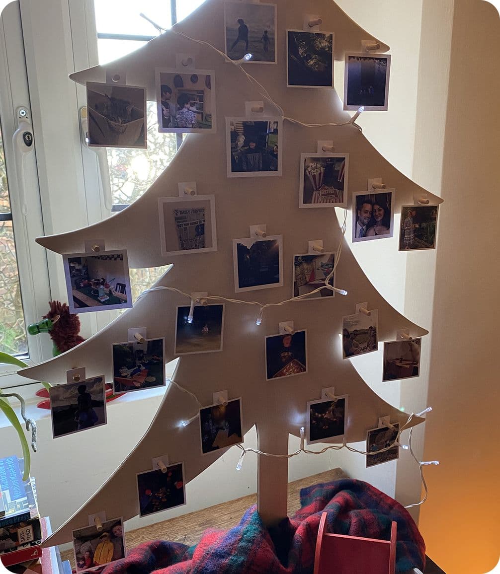 Cardboard tree with polaroid photos clipped to it, adorned with string lights, set by a window with a red plaid blanket at the base.