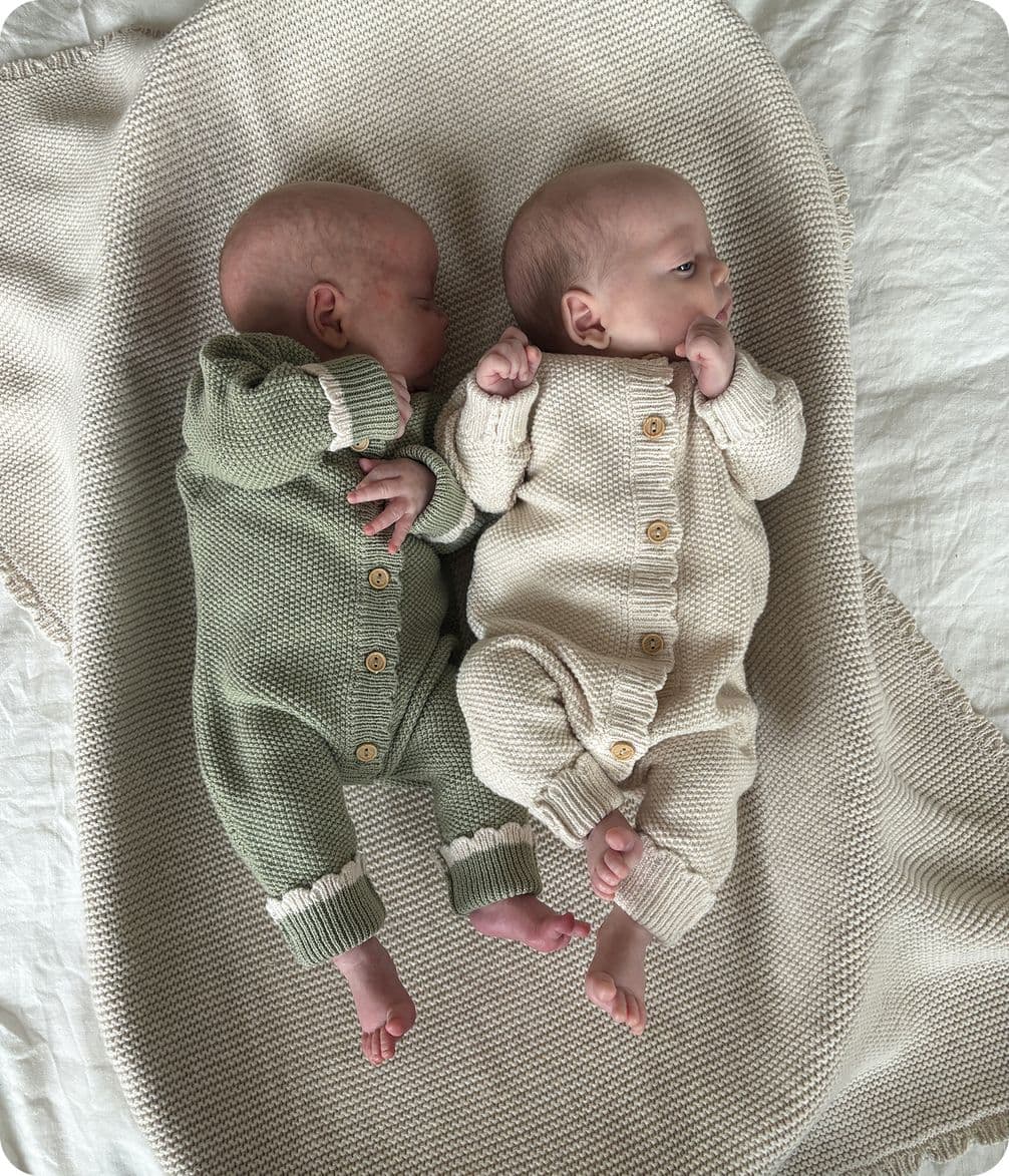Two babies lie on a soft, textured blanket. One wears a green knit outfit, the other a cream one, both with button details.