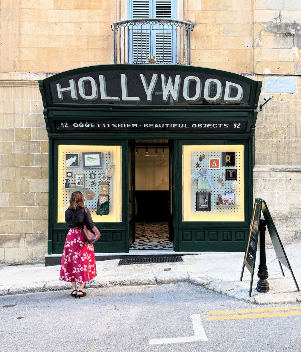 Woman in a red floral skirt stands outside a shop with "HOLLYWOOD" sign. The display features various objects and artwork.