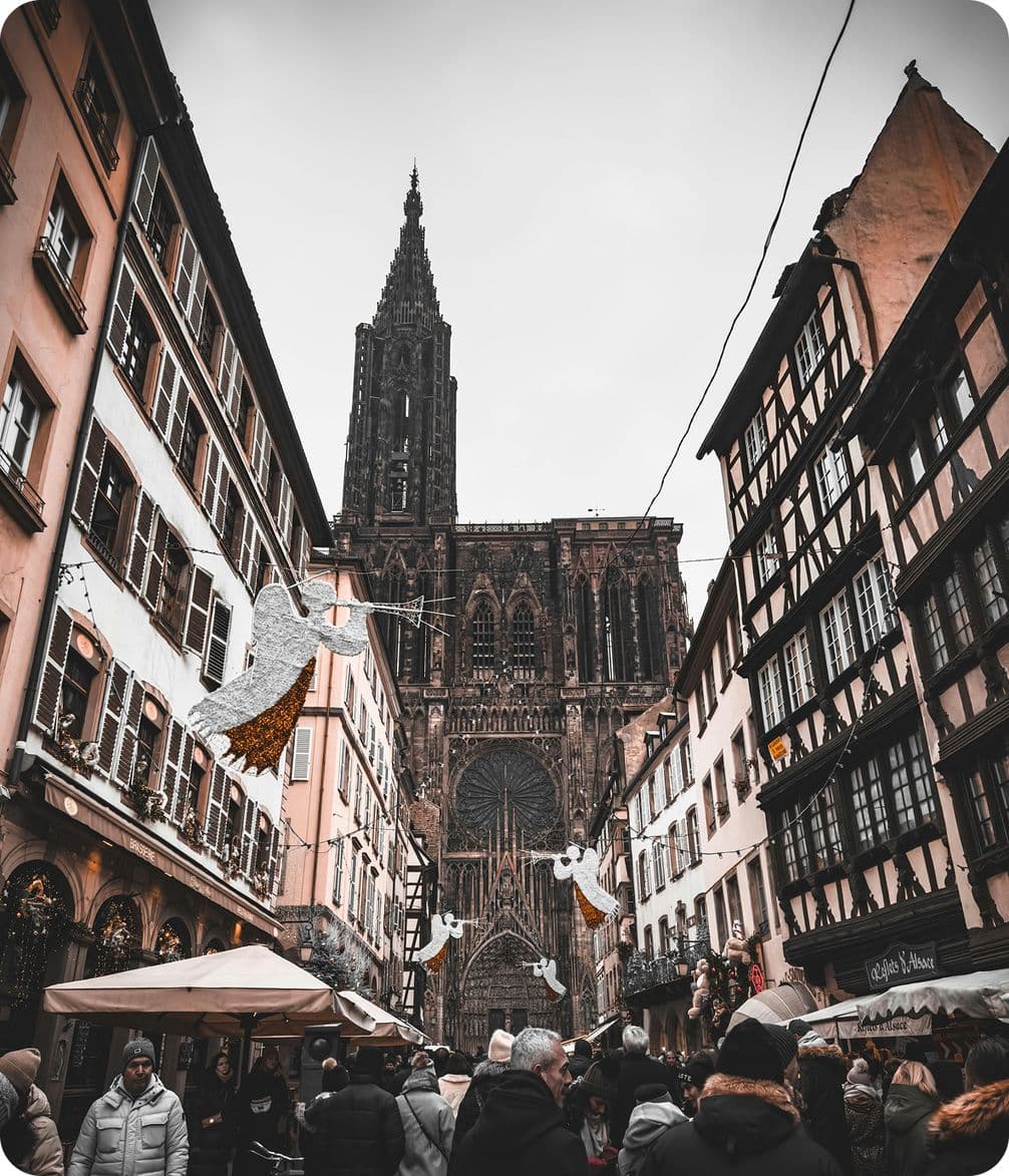 Crowded street with festive decorations, leading to a large Gothic cathedral. Historic buildings line the street, creating a charming atmosphere.