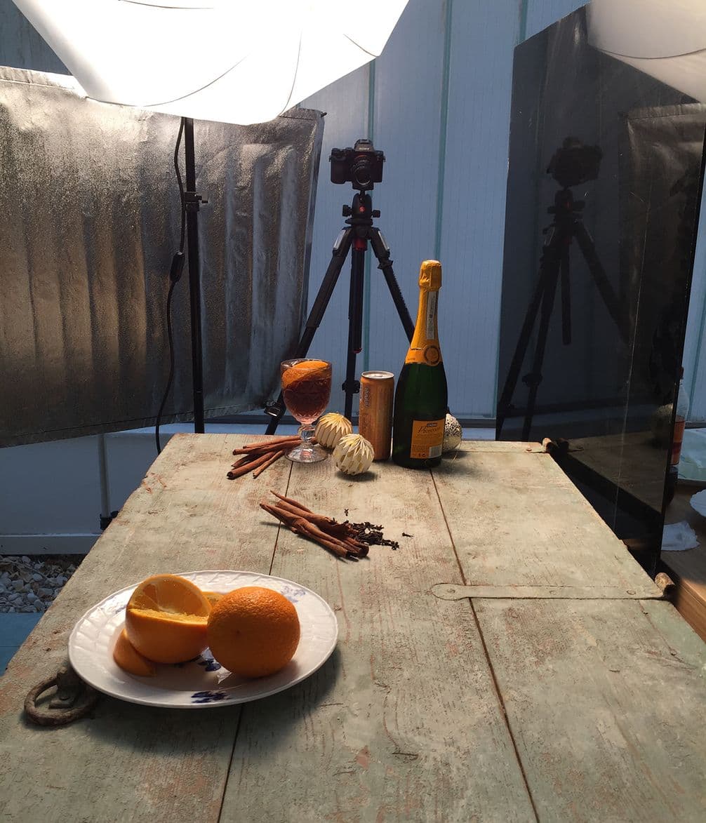 A rustic table with oranges, cinnamon, cloves, and a bottle of champagne arranged for a photoshoot. Cameras and lights in the background.