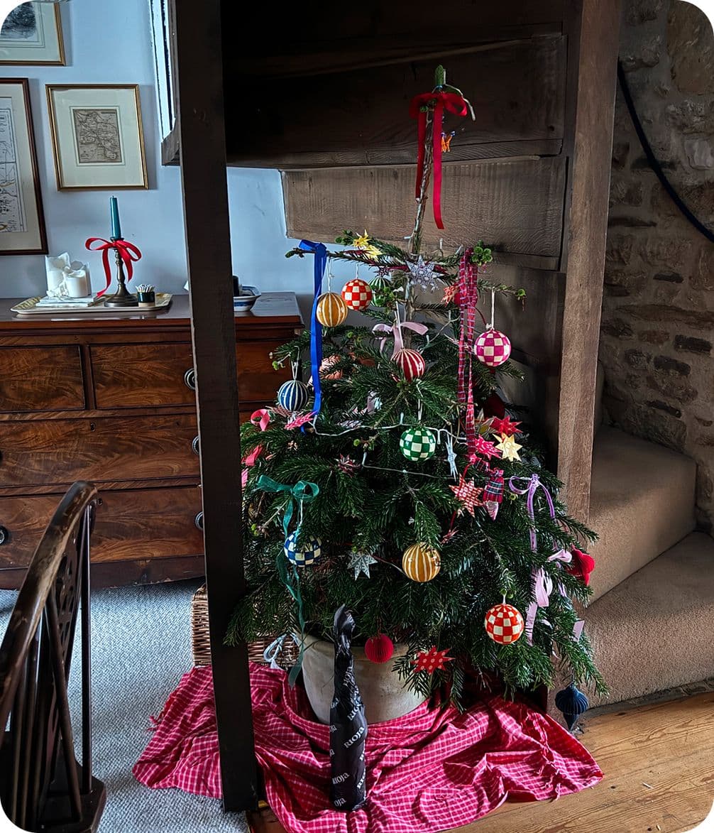 Small Christmas tree with colorful ornaments and ribbons, placed on a red checkered cloth near a wooden staircase and dresser.