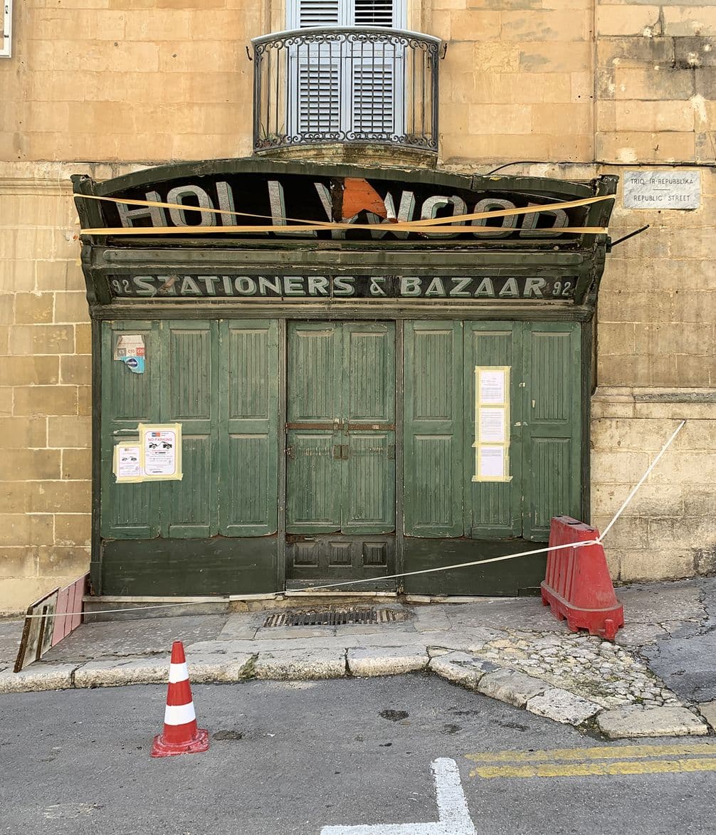 Old green storefront with "Hollywood Stationers & Bazaar" sign, boarded windows, and construction cones on a narrow cobblestone street.