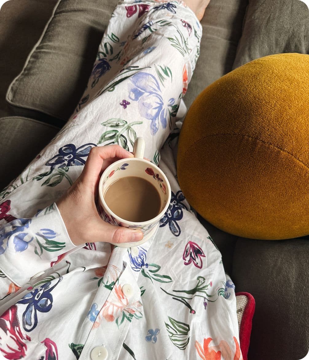 Person in floral pajamas holding a mug of coffee, sitting on a couch with a mustard-yellow pillow beside them.