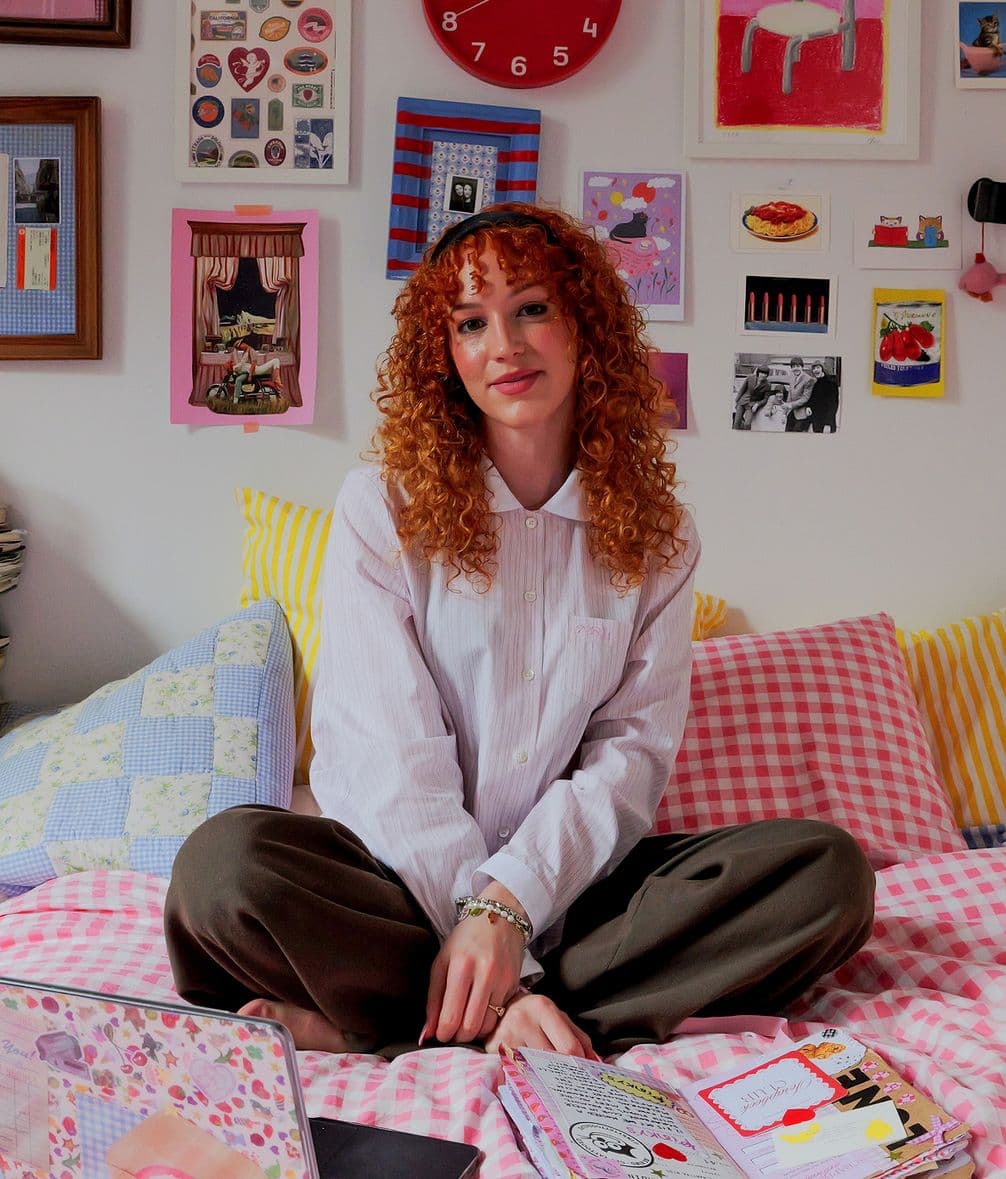 Smiling curly-haired woman seated cross-legged on a patterned bed surrounded by colorful pillows, wall art, a laptop and notebooks.