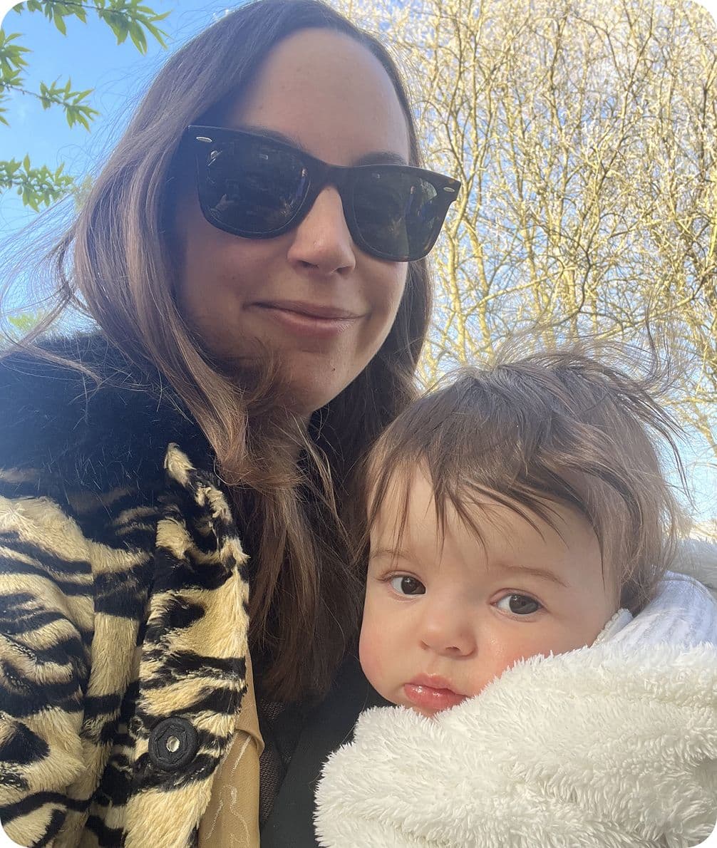 Woman in sunglasses holding a baby wrapped in a white blanket, outdoors with trees in the background.