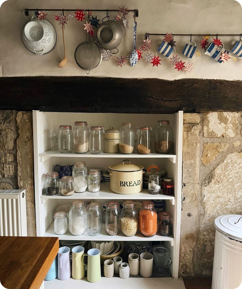 A rustic kitchen shelf with jars of grains, a bread tin, and mugs. Decorative stars and kitchenware hang above on a wooden beam.