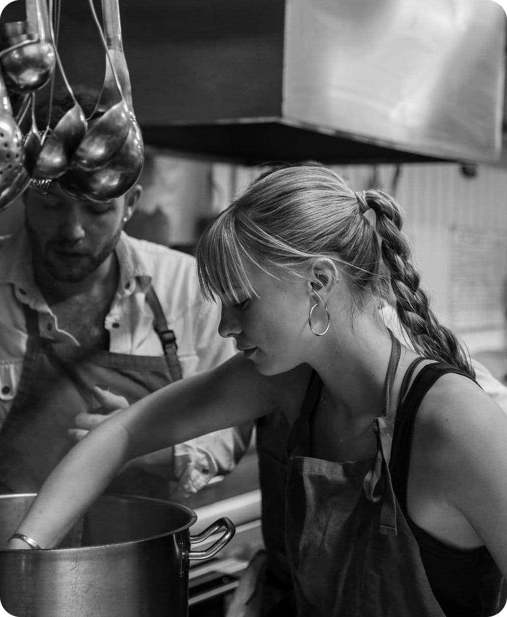 Black-and-white kitchen scene: woman with braided hair stirs a large pot; male cook watches, metal ladles hanging above.