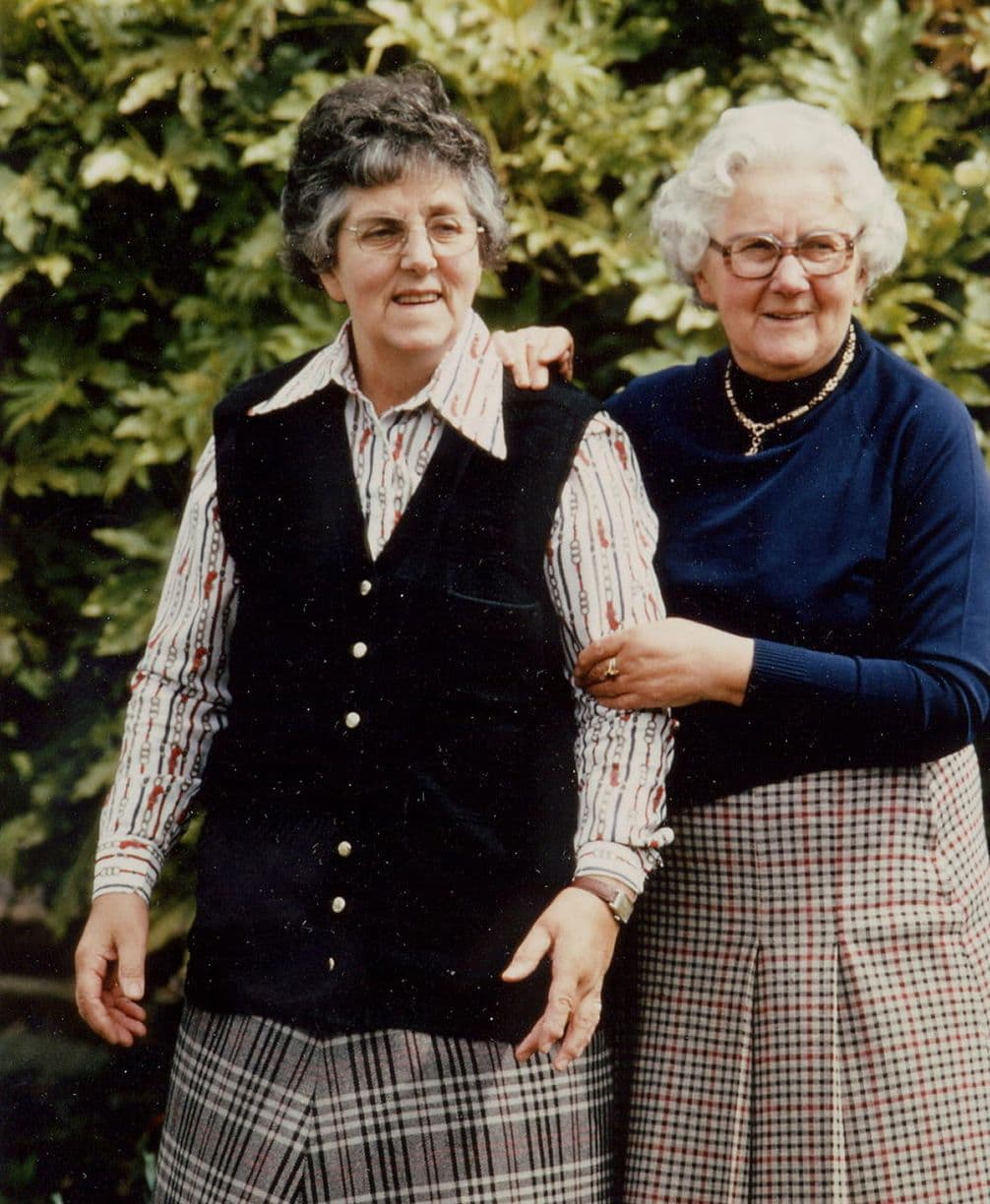 Two elderly women standing together, smiling, in front of leafy bushes. One wears a black vest; the other, glasses and a navy sweater.