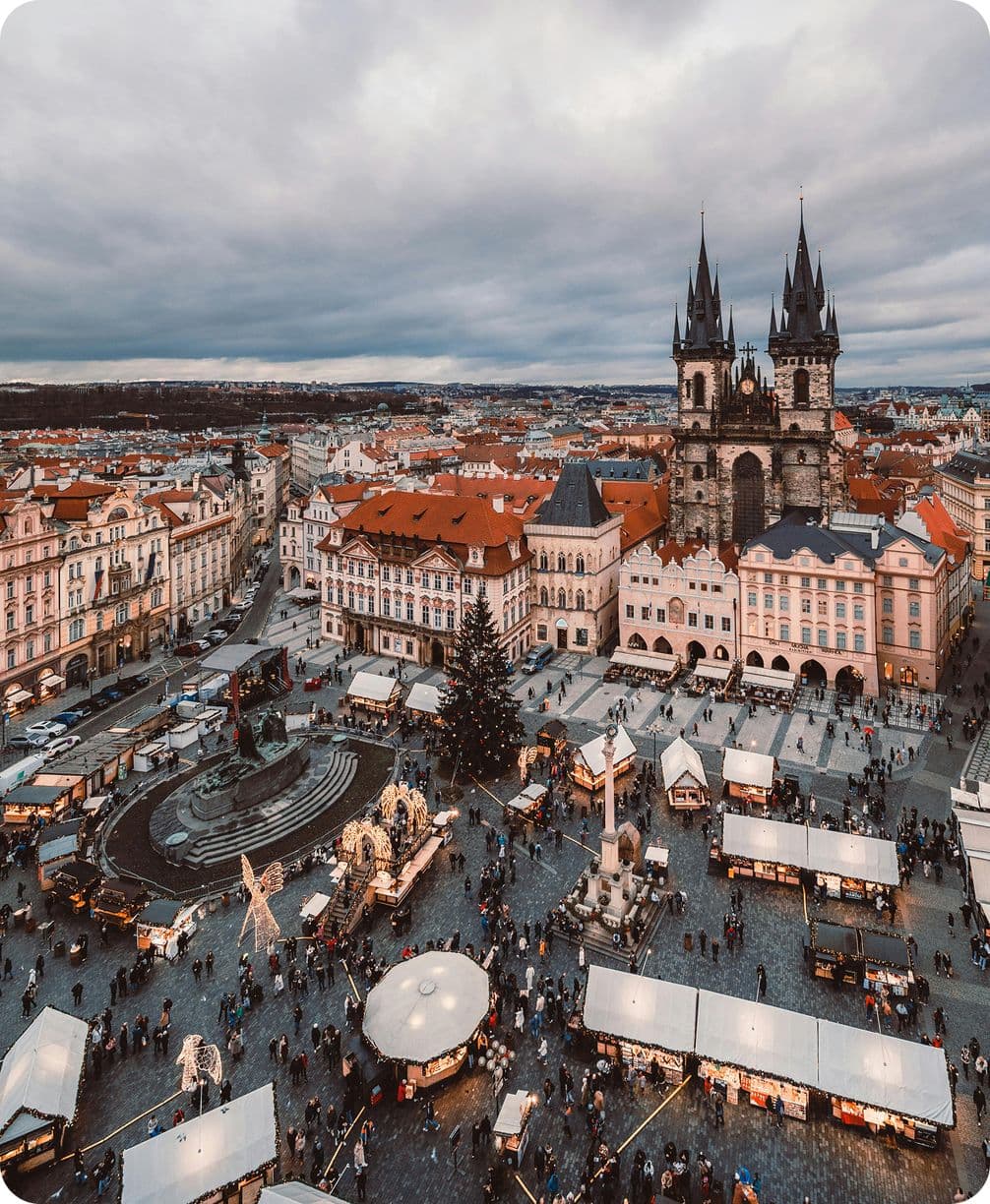 Aerial view of a bustling Prague with a Christmas market, historic buildings, and a prominent Gothic church under a cloudy sky.