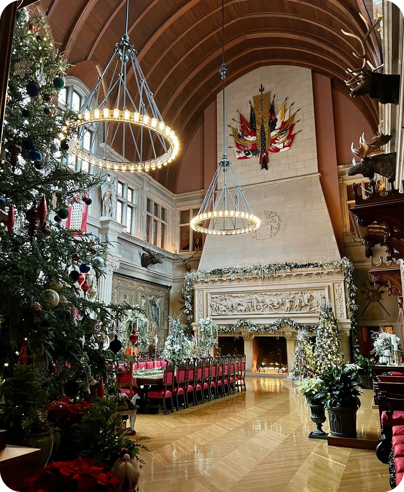 A grand hall decorated for Christmas with a tall tree, long dining table, ornate fireplace, and large chandeliers under a vaulted ceiling.