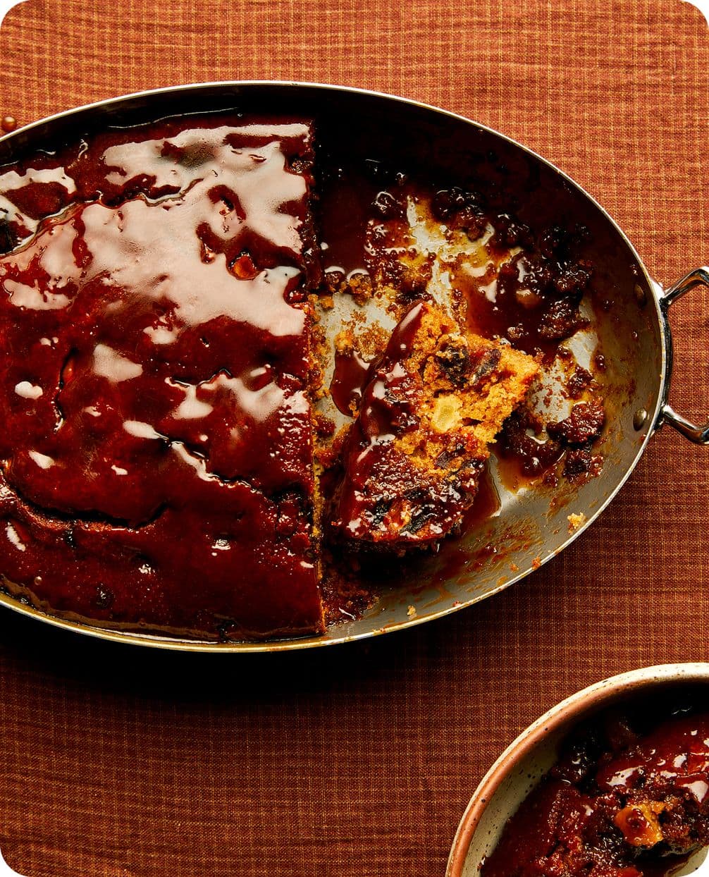 Oval pan of glazed meatloaf with a slice removed, showing crumbly interior and rich brown sauce on an orange tablecloth.