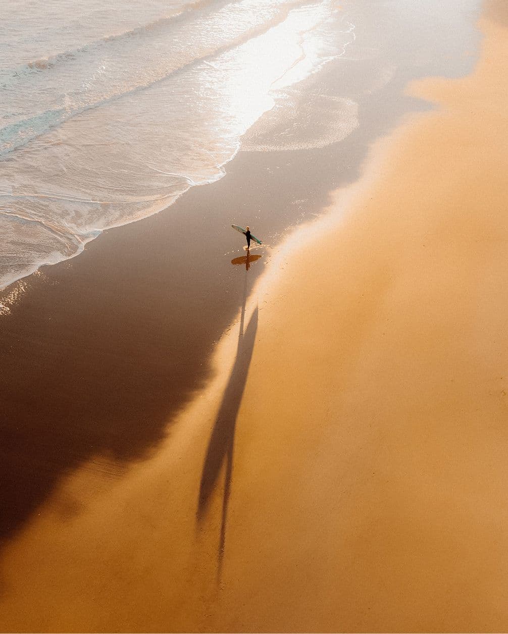 Drone photograph of a surfer approaching the water from a sandy beach