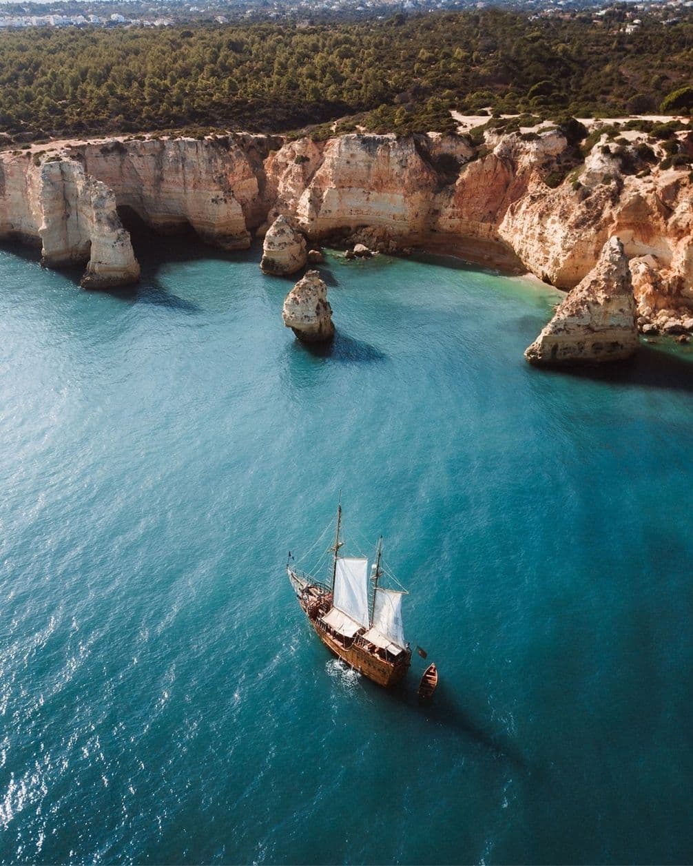 Aerial view of a sailing ship near rugged cliffs and turquoise waters, with lush greenery atop the cliffs under a clear sky.